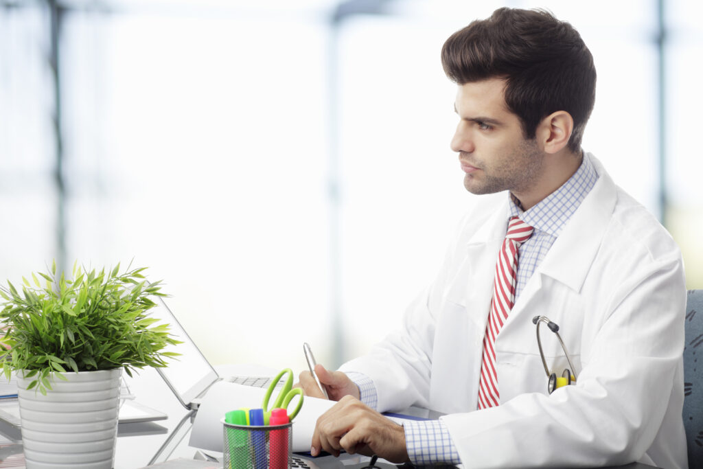 Close-up of a young male medical professional in a lab coat and tie using a laptop, representing the career growth and how much do medical billing and coding make as specialists.