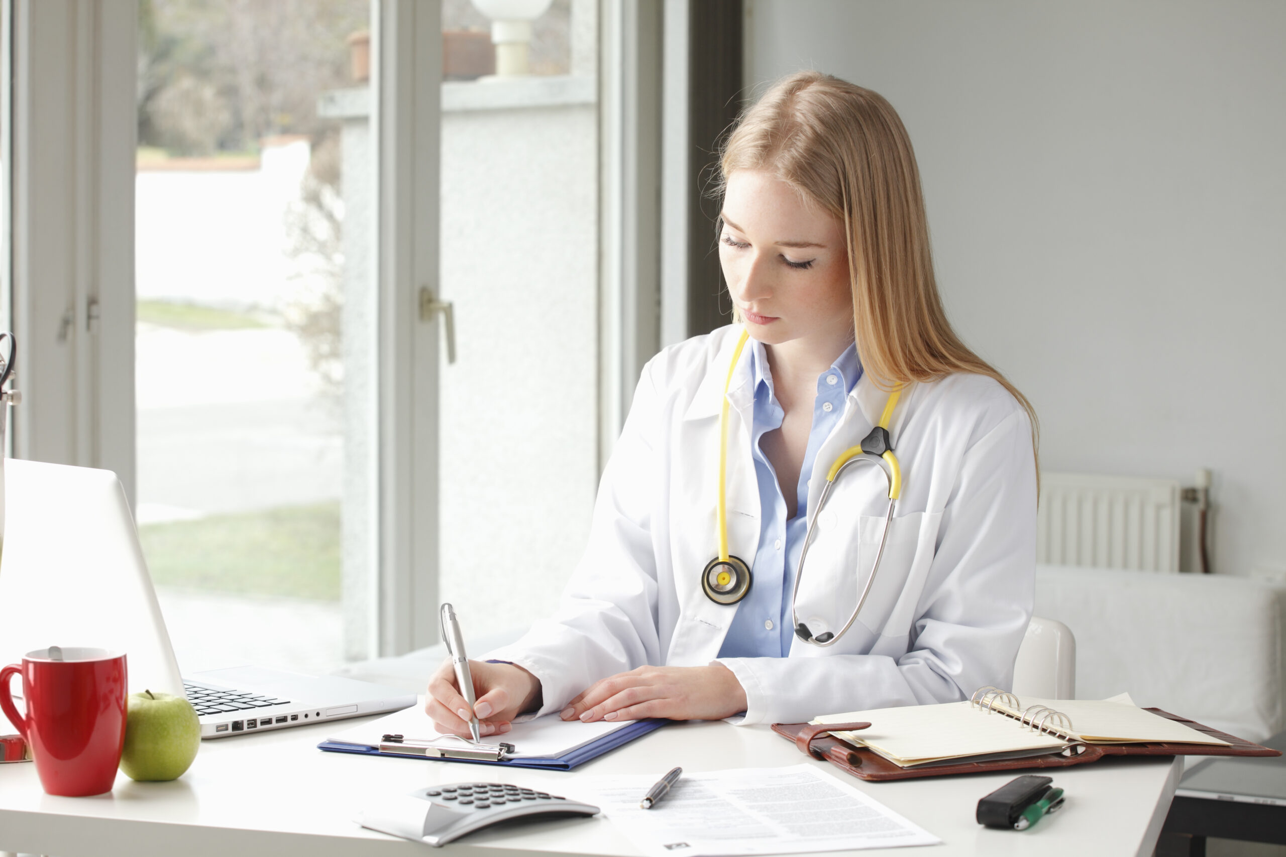A female doctor with a stethoscope sits at a clean white desk, meticulously writing on a clipboard, symbolizing the human audit process within a high-tier medical billing service.