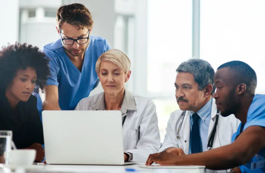 A diverse medical team collaborates in front of a laptop, demonstrating the expert human oversight required for artificial intelligence in revenue cycle management to prevent clinical errors.